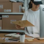 Wanita menyortir paket di meja kerja kantor logistik, mencerminkan aktivitas harian agen pengiriman barang
