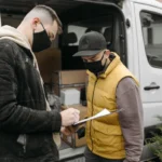 Dua orang bertanda tangan pada dokumen tanda terima barang di samping kendaraan pengiriman